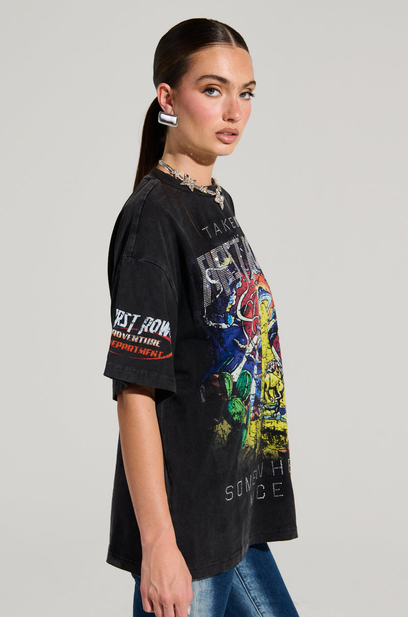 A woman with a long ponytail wearing a FIRST ROW MIDNIGHT RHINESTONE EMBELLISHED T-SHIRT and blue jeans stands against a plain background, showcasing colorful graphics and rhinestone lettering, as she listens to her earphones, glancing left.