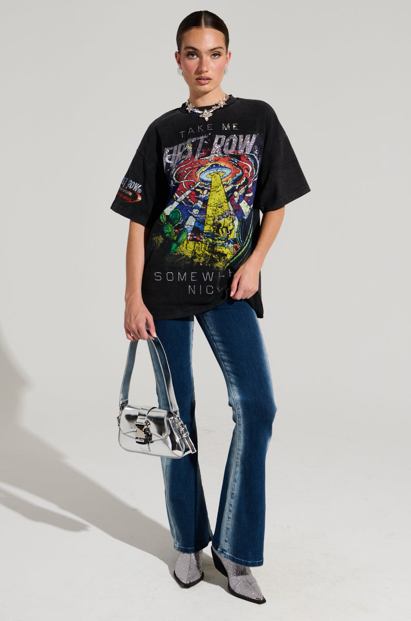 Someone stands against a plain background wearing a black FIRST ROW MIDNIGHT RHINESTONE EMBELLISHED T-SHIRT, blue jeans, and silver ankle boots. They accessorize with a small metallic handbag, a chunky necklace, and have their hair pulled back.