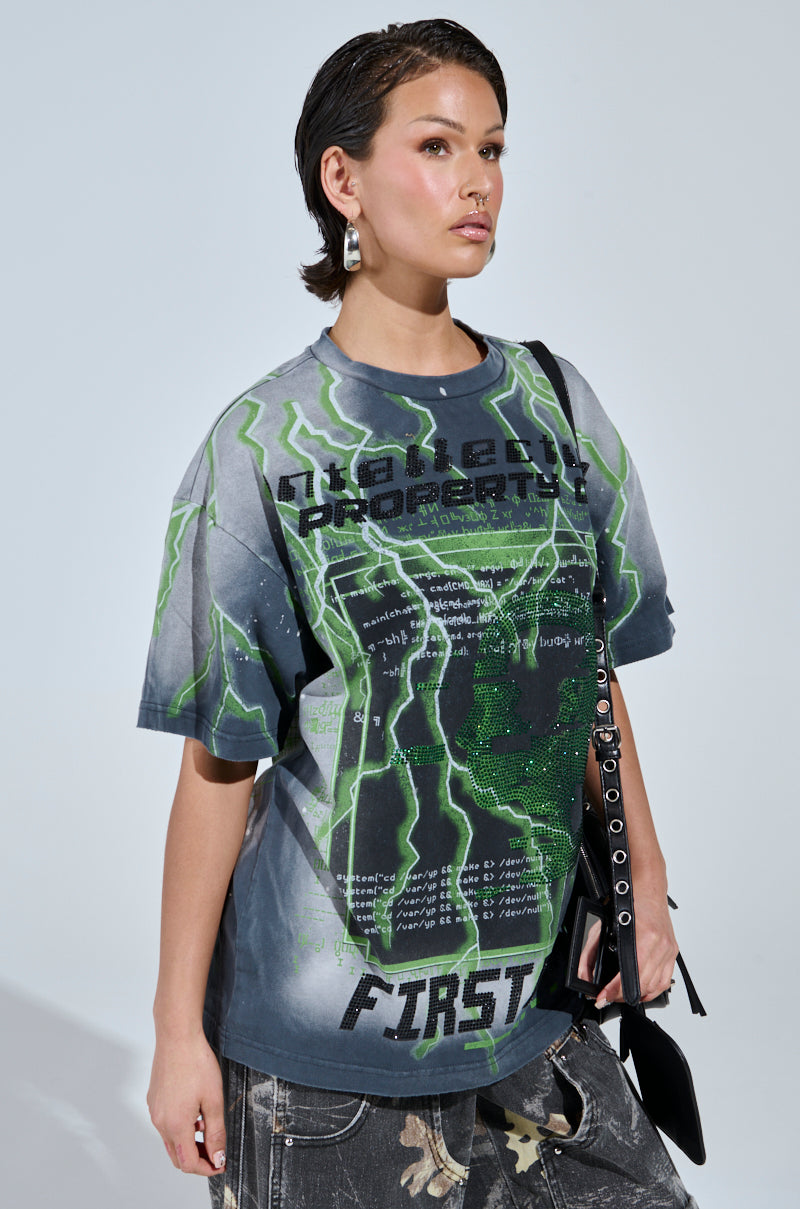 Wearing the FIRST ONE HERE OVERSIZED GRAPHIC TEE with a futuristic design, they pair it with dark patterned pants. Their short hair and black purse enhance the look against a plain, light-colored background.