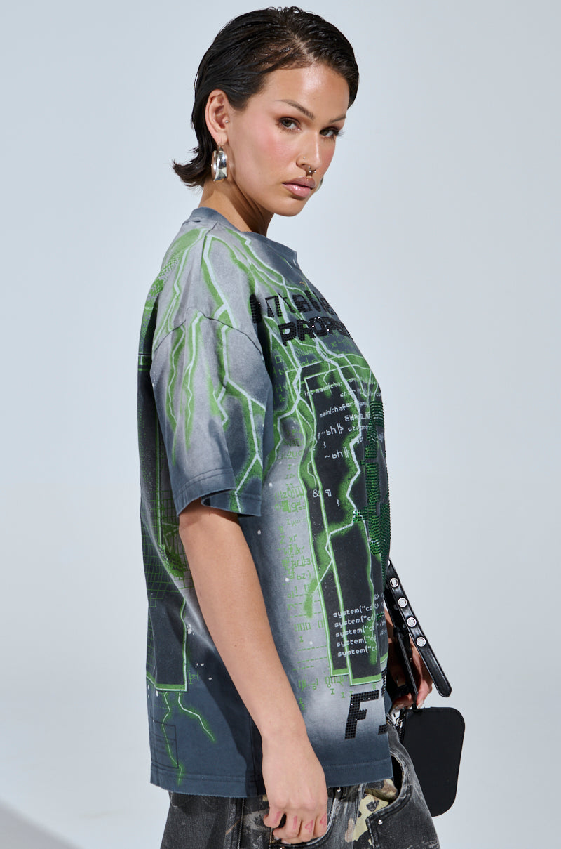 A person with short, dark hair wears the FIRST ONE HERE OVERSIZED GRAPHIC TEE in gray and green featuring a futuristic design. They're holding a black object, wearing silver hoop earrings, and posing sideways against a light background.