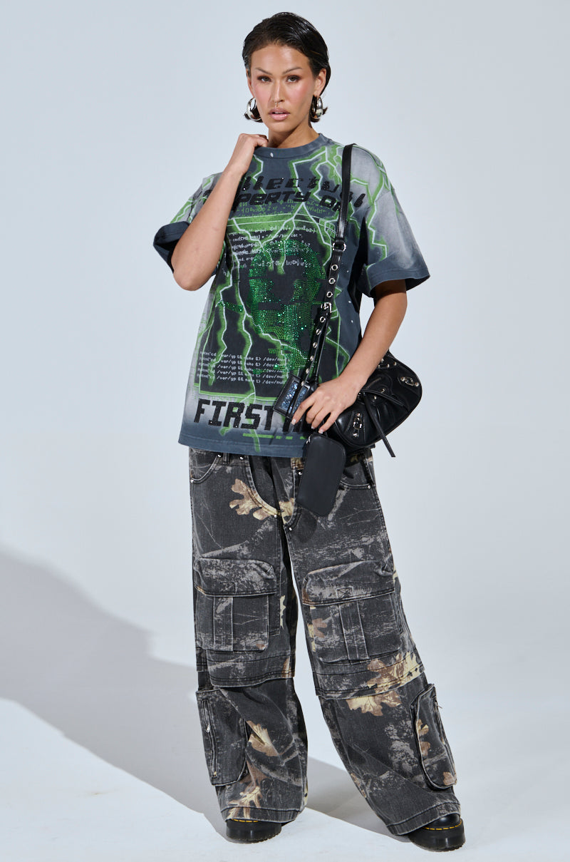 Wearing the FIRST ONE HERE OVERSIZED GRAPHIC TEE with a green lightning design, a person pairs it with wide-legged, patterned cargo pants and carries a black shoulder bag against a plain backdrop, highlighting their eclectic style and futuristic graphic print flair.