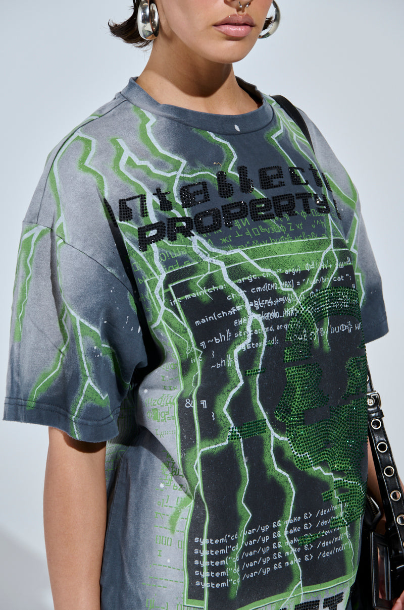 A person wears the FIRST ONE HERE OVERSIZED GRAPHIC TEE, featuring a gray base with a green lightning design and "Intellectual Property" text adorned with computer code. With short hair and carrying a black bag with a chain strap, they exude an edgy, futuristic style.