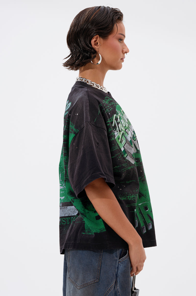A person with short, dark wet-look hair wears the PROPERTY OVERSIZED GRAPHIC TEE featuring green designs, paired with blue jeans, a chunky chain necklace, and large hoop earrings, standing in profile against a plain white background.