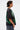 A person with short, dark wet-look hair wears the PROPERTY OVERSIZED GRAPHIC TEE featuring green designs, paired with blue jeans, a chunky chain necklace, and large hoop earrings, standing in profile against a plain white background.