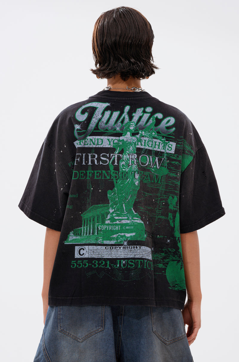 A person with short, dark hair wears the PROPERTY OVERSIZED GRAPHIC TEE, a black lightweight cotton shirt featuring green graphics, white “Justice” and “First Row” text on the back, Statue of Liberty imagery, and a phone number.