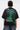 A person with short, dark hair wears the PROPERTY OVERSIZED GRAPHIC TEE, a black lightweight cotton shirt featuring green graphics, white “Justice” and “First Row” text on the back, Statue of Liberty imagery, and a phone number.