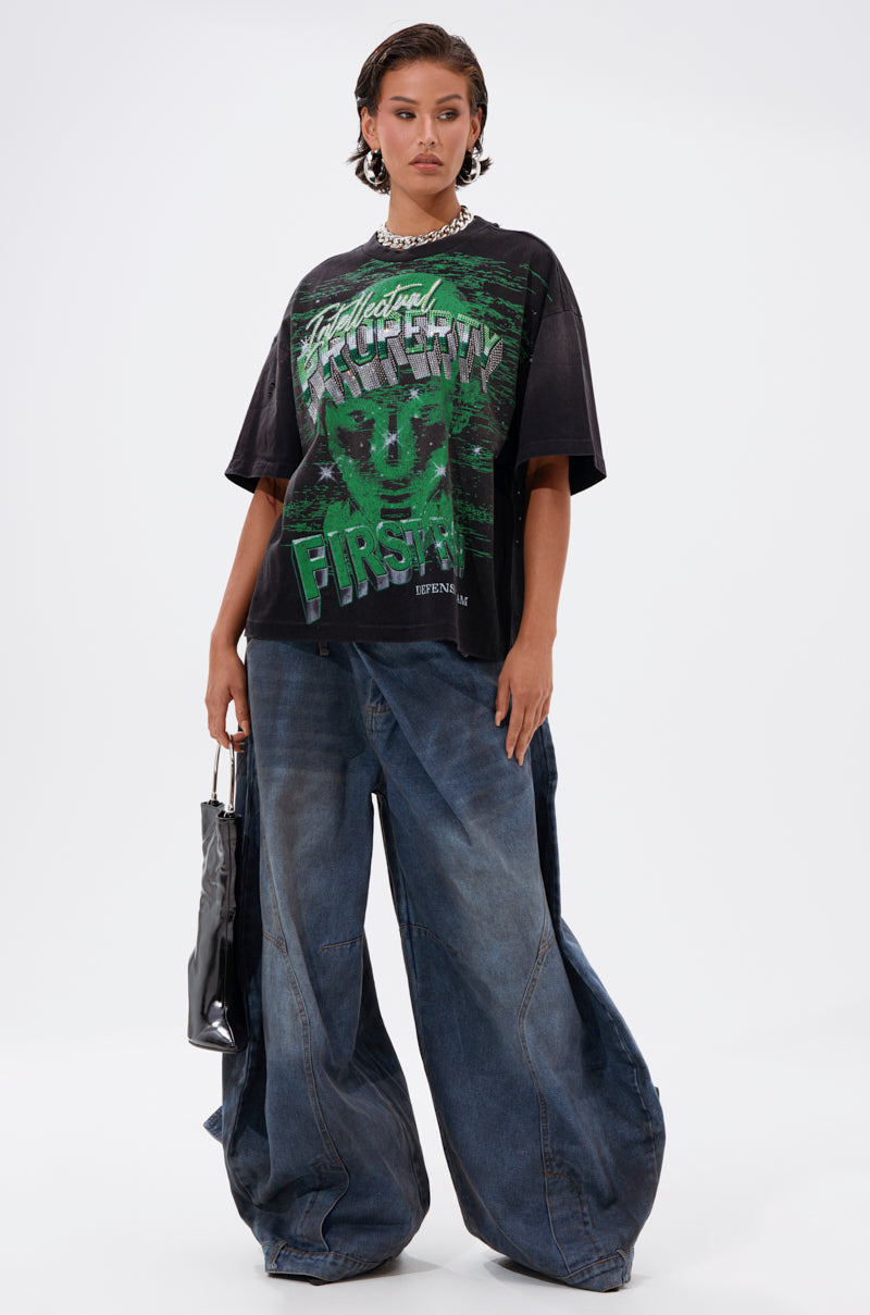 A person stands against a plain white background, confidently holding a shiny black bag and looking to the side while wearing the PROPERTY OVERSIZED GRAPHIC TEE and extremely wide-leg blue jeans.