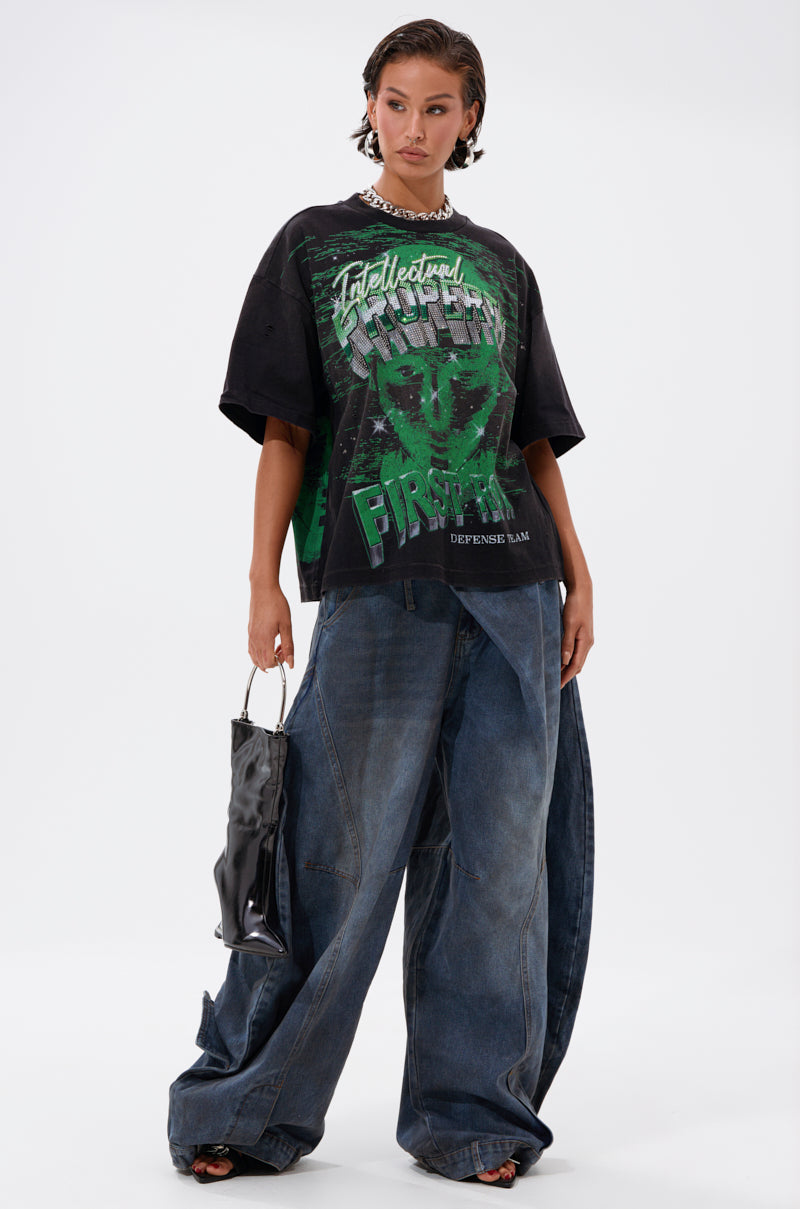 A woman poses in the PROPERTY OVERSIZED GRAPHIC TEE with green lettering, paired with wide-leg blue jeans, chunky shoes, a shiny black handbag, chain necklace, and slicked-back short hair.