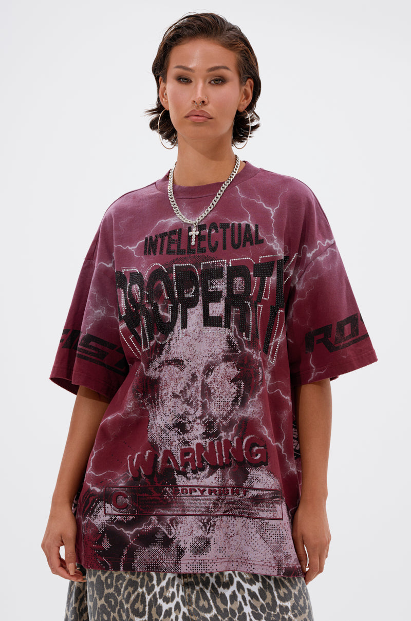 A person with short, wavy hair wears the INTELLECTUAL PROPERTY GRAPHIC TEE IN BURGUNDY, featuring bold graphics and a warning motif, paired with a leopard print skirt.