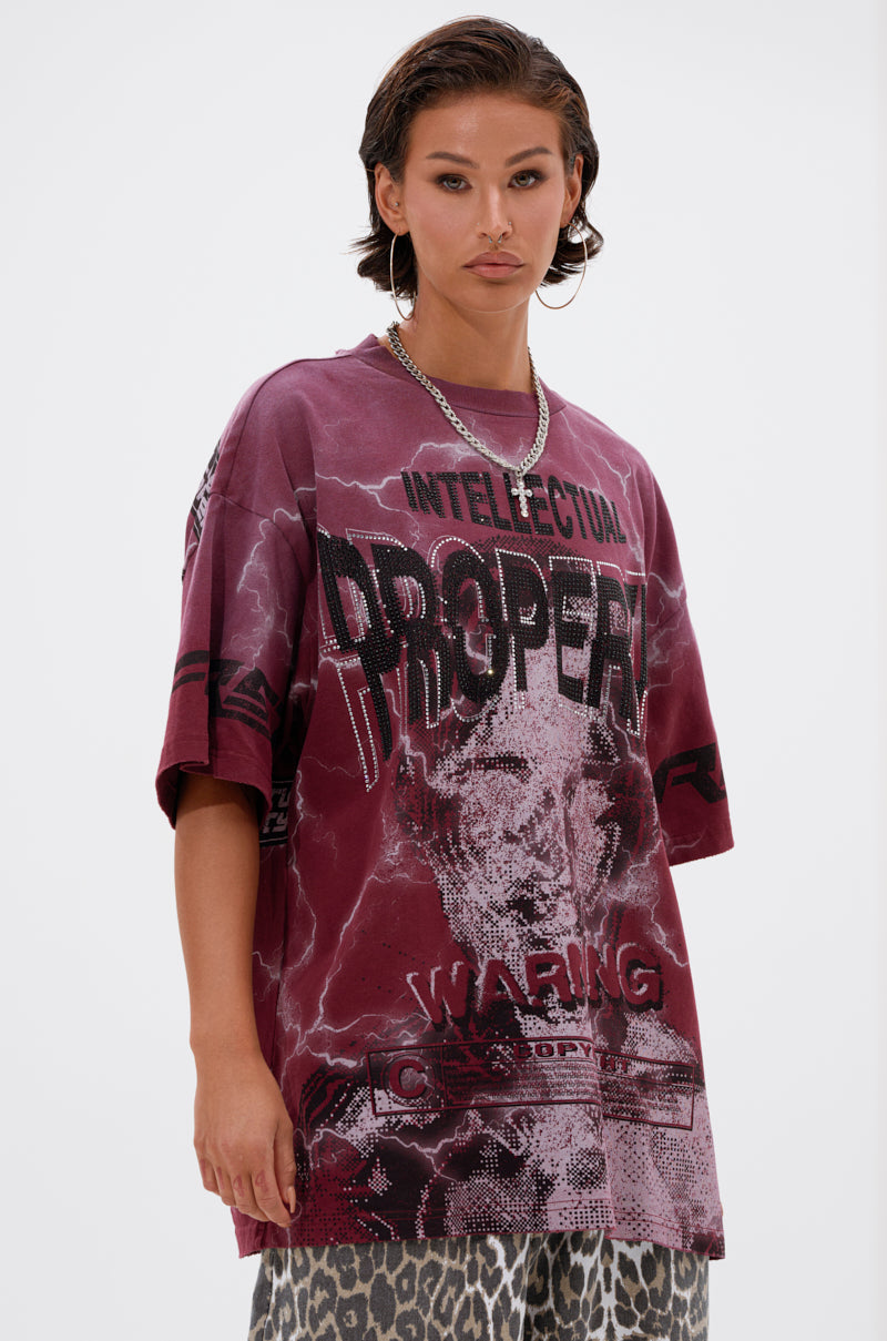 A person with short, slicked-back hair models the oversized INTELLECTUAL PROPERTY GRAPHIC TEE IN BURGUNDY, featuring bold text and an abstract face, styled with grey leopard-print pants against a plain light backdrop.