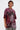 A person with short, slicked-back hair models the oversized INTELLECTUAL PROPERTY GRAPHIC TEE IN BURGUNDY, featuring bold text and an abstract face, styled with grey leopard-print pants against a plain light backdrop.