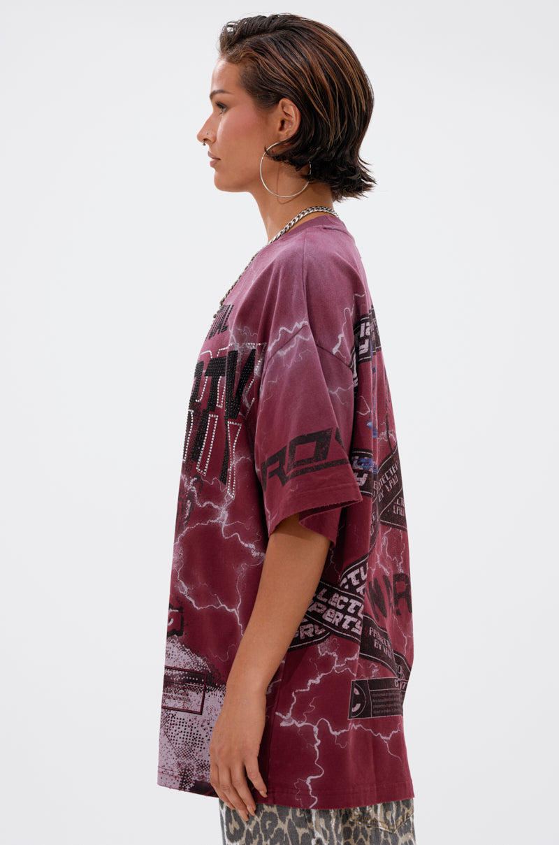 A person with short dark hair and large hoop earrings wears the INTELLECTUAL PROPERTY GRAPHIC TEE IN BURGUNDY and leopard print pants, standing in profile against a plain light background.