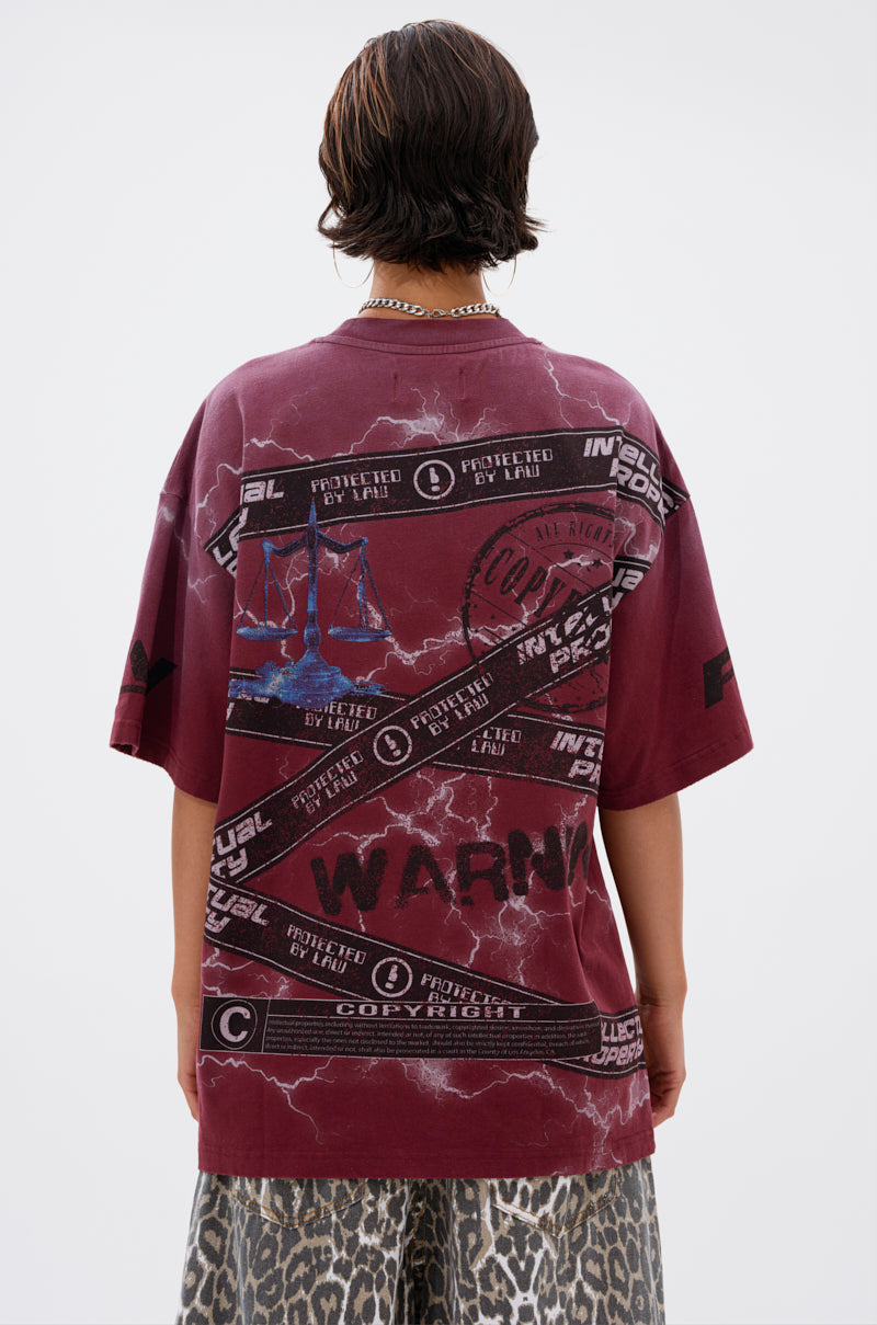 A person with short brown hair, seen from behind, wears the INTELLECTUAL PROPERTY GRAPHIC TEE IN BURGUNDY featuring bold warning text and copyright symbols, paired with leopard print pants against a plain light background.