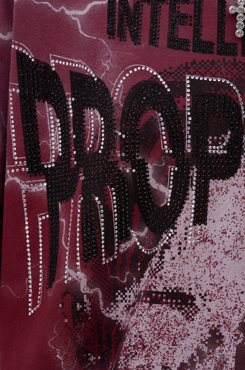 Close-up of burgundy fabric from the INTELLECTUAL PROPERTY GRAPHIC TEE IN BURGUNDY, featuring black beaded and rhinestone "DROP" lettering with partial letters above on a textured, abstract pattern background.
