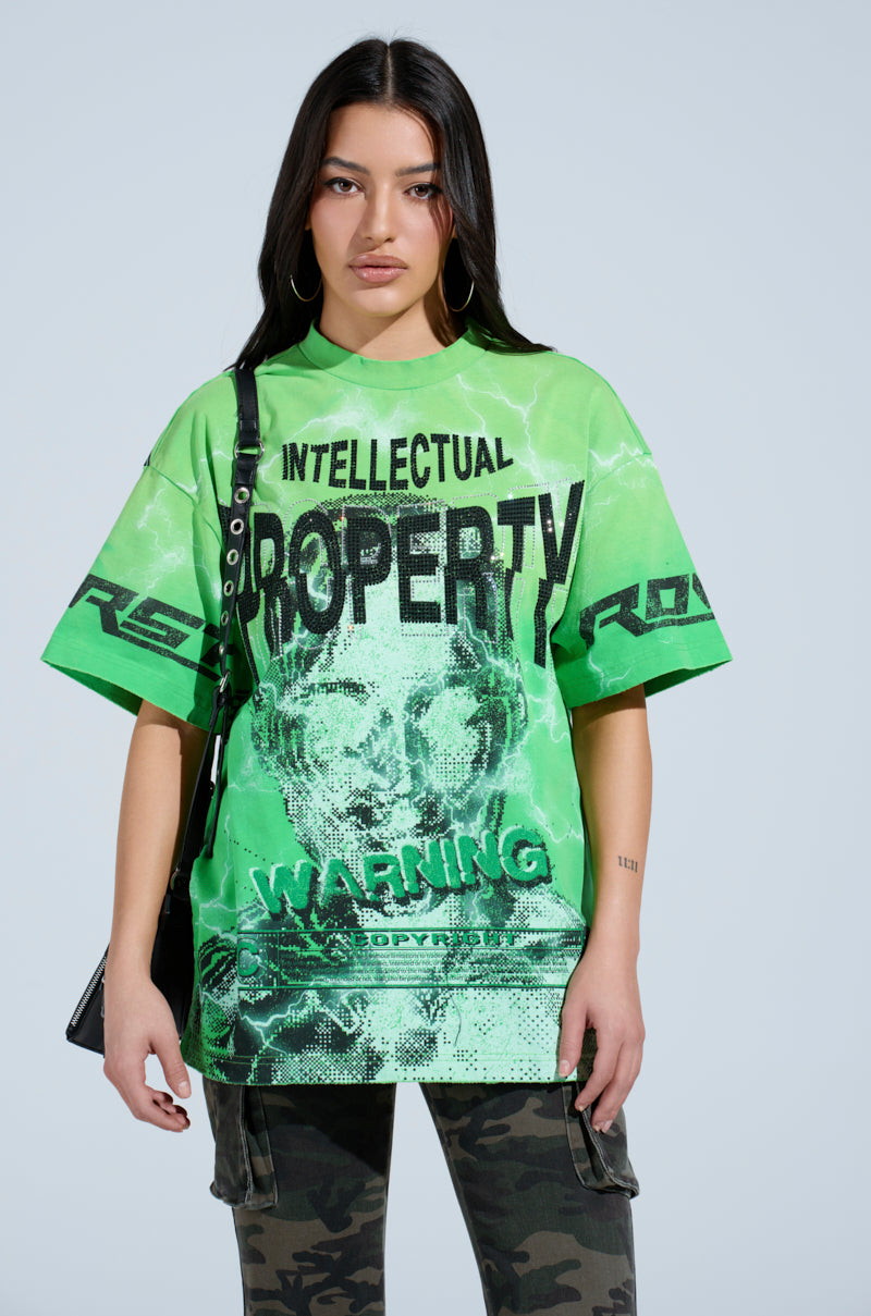 Wearing an oversized INTELLECTUAL PROPERTY GRAPHIC TEE in green, featuring bold "INTELLECTUAL PROPERTY" and "WARNING" graphics, the woman with long dark hair and hoop earrings carries a black strap. Her look is stylishly completed with camo pants for a striking ensemble.