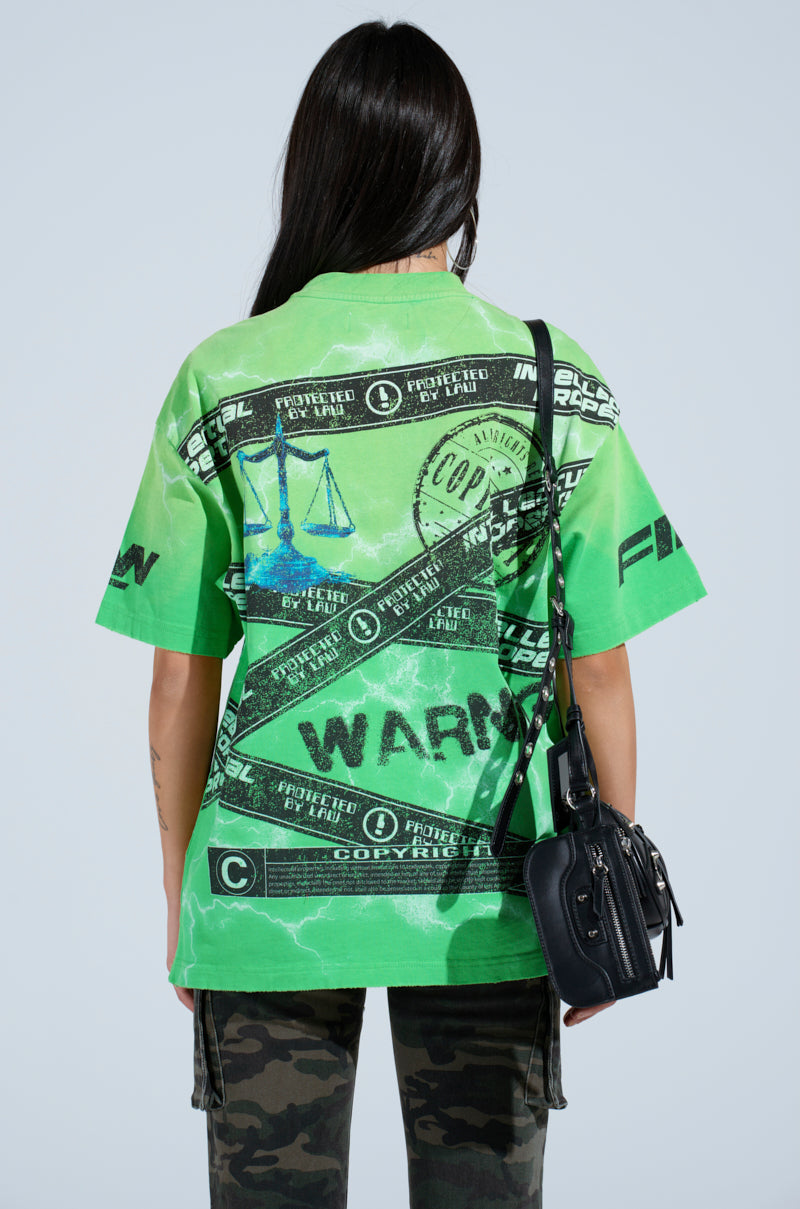 A person with long hair dons an oversized silhouette featuring a green INTELLECTUAL PROPERTY GRAPHIC TEE, embellished with striking scales of justice and copyright symbols. Camouflage pants complete the look, paired with a black handbag against a plain, light-toned backdrop.