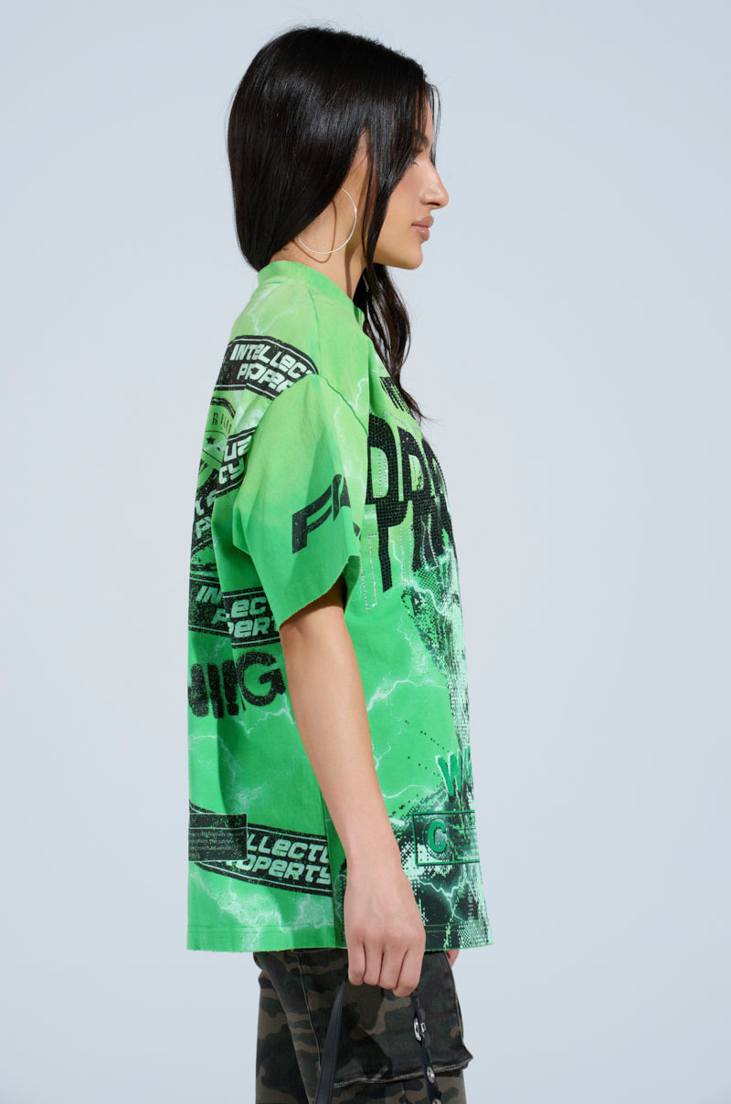 A woman with long dark hair wears a vibrant green Intellectual Property Graphic Tee, featuring sports-themed prints, and camo-patterned pants. She's standing sideways against a plain backdrop while holding a bag.
