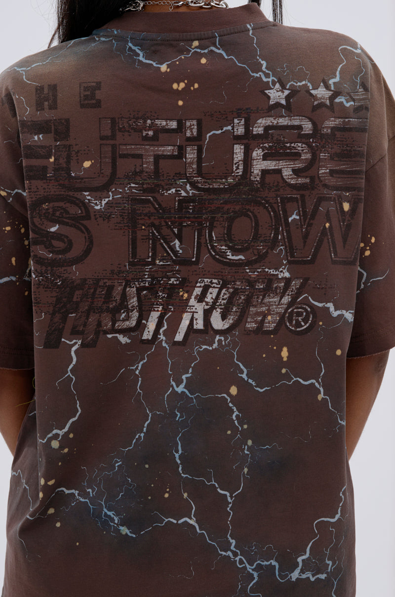 Someone wearing the HUMANITY OVERSIZED GRAPHIC TEE in brown, which features a futuristic lightning design and bold, distressed "THE FUTURE IS NOW FIRST ROW®" text on the back, with white and yellow speckled accents. 7