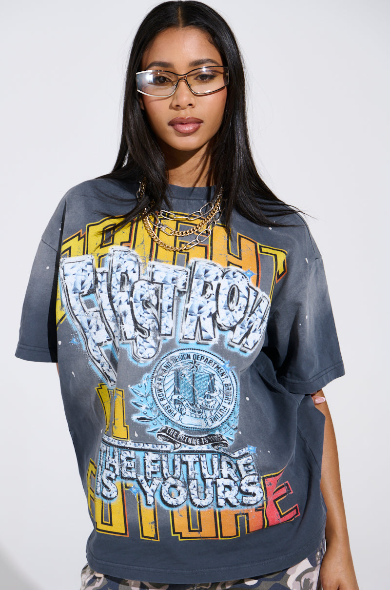 Against a plain backdrop, a woman with long hair showcases large, clear glasses and an oversized cityscape print tee named "THE FUTURE IS YOURS." She adds flair to her look with layered gold necklaces.
