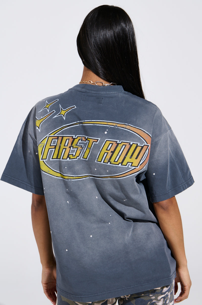 Wearing "THE FUTURE IS YOURS" oversized gray graphic tee with a vibrant "First Row" design, star graphics, and rhinestone embellishments, a long-haired person pairs it with camouflage pants against a light-colored backdrop.