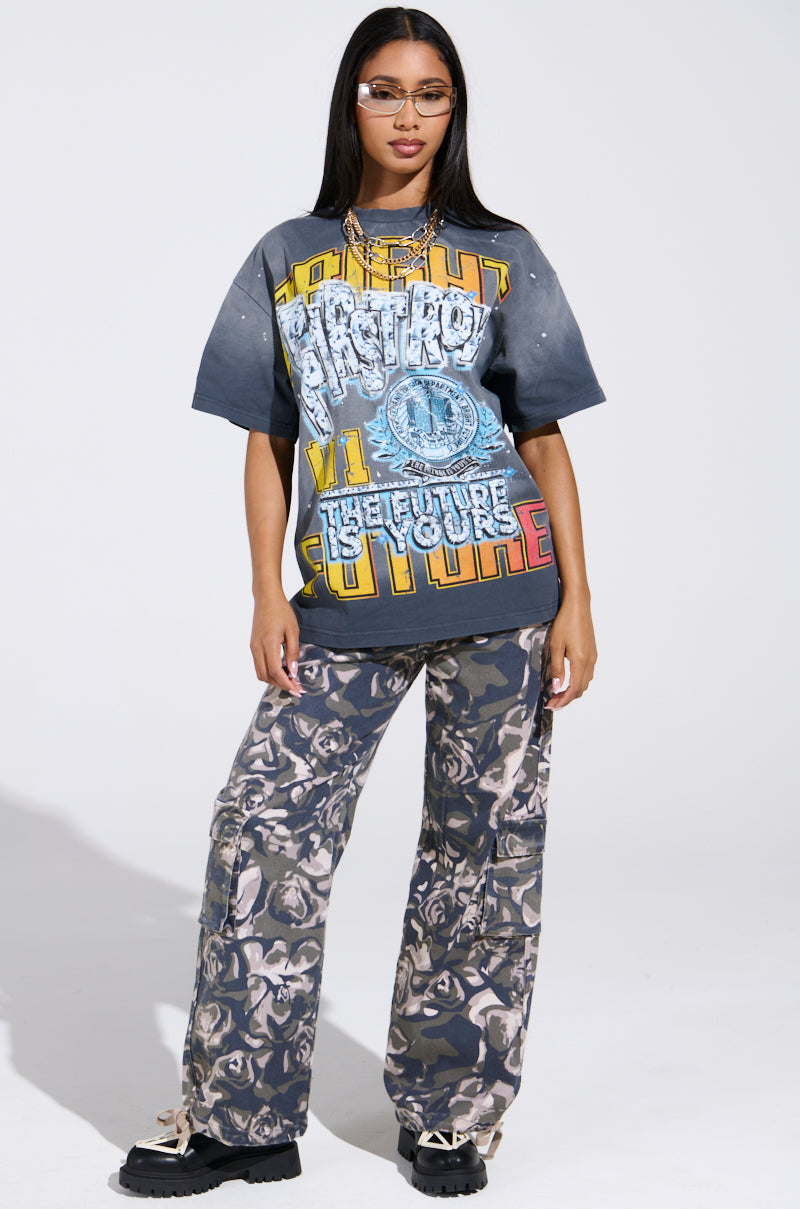 Wearing "THE FUTURE IS YOURS" oversized graphic tee with vibrant text and rhinestones, paired with camo pants and black sneakers, a long-haired person with glasses and a necklace stands confidently against a plain background.