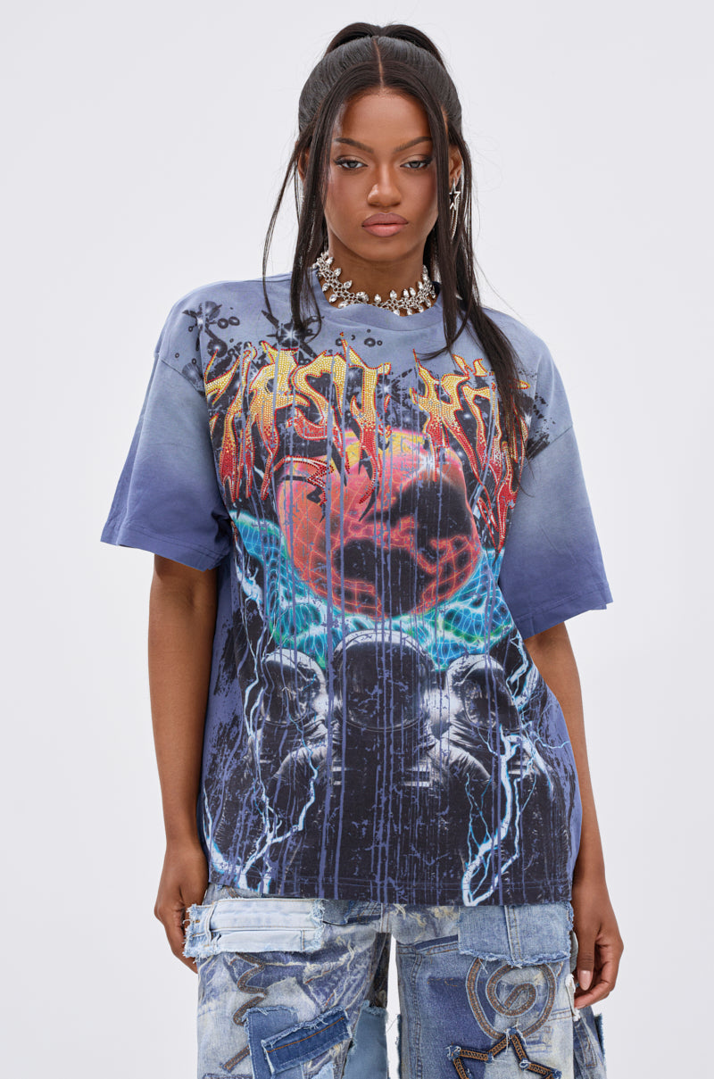 A woman with long, dark pigtails wears the IRREVERENT OVERSIZED GRAPHIC TEE with lightning, blue and red graphics, and flames, paired with distressed patch jeans. She stands against a plain light background.