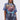 A woman with long, dark pigtails wears the IRREVERENT OVERSIZED GRAPHIC TEE with lightning, blue and red graphics, and flames, paired with distressed patch jeans. She stands against a plain light background.