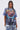 A woman with long, dark pigtails wears the IRREVERENT OVERSIZED GRAPHIC TEE with lightning, blue and red graphics, and flames, paired with distressed patch jeans. She stands against a plain light background.