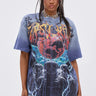 A woman with long, dark pigtails wears the IRREVERENT OVERSIZED GRAPHIC TEE with lightning, blue and red graphics, and flames, paired with distressed patch jeans. She stands against a plain light background.