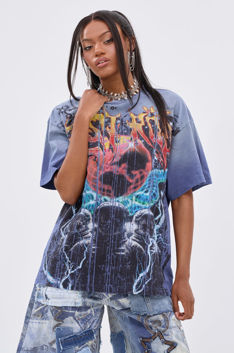 A woman with long straight hair wears the IRREVERENT OVERSIZED GRAPHIC TEE featuring a bold lightning graphic, paired with patchwork denim jeans. She accessorizes with layered necklaces and hoop earrings, standing against a plain white background.