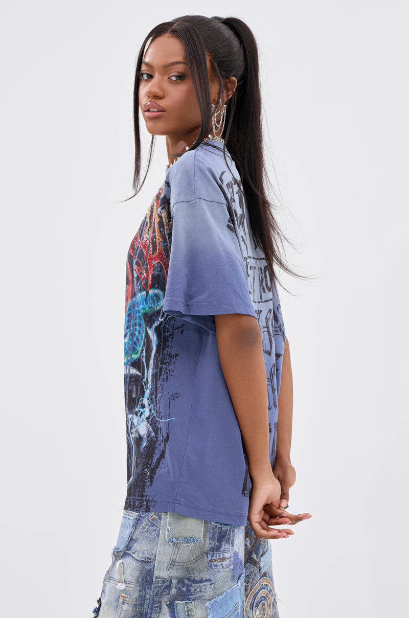 A woman with long dark hair in a high ponytail wears the IRREVERENT OVERSIZED GRAPHIC TEE and patchwork denim jeans. She stands sideways with her hands behind her back, looking toward the camera against a plain light background.
