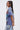 A woman with long dark hair in a high ponytail wears the IRREVERENT OVERSIZED GRAPHIC TEE and patchwork denim jeans. She stands sideways with her hands behind her back, looking toward the camera against a plain light background.