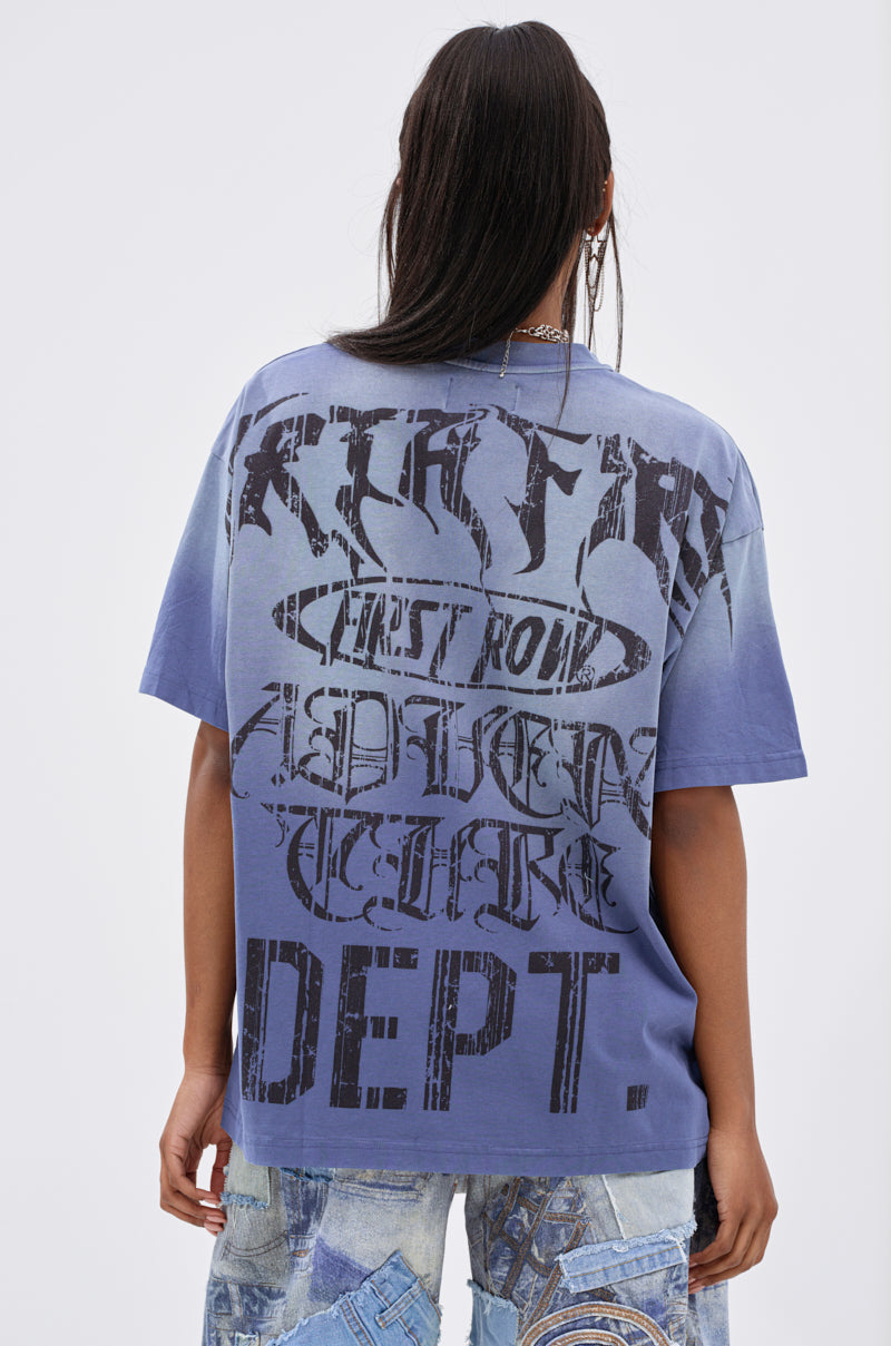 A person with long dark hair stands facing away, wearing the IRREVERENT OVERSIZED GRAPHIC TEE with bold black lettering and a lightning graphic on the back, paired with patchwork denim jeans.