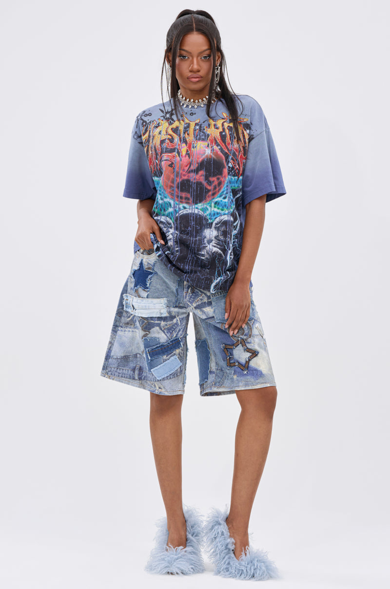 A person stands facing forward in the IRREVERENT OVERSIZED GRAPHIC TEE, paired with patchwork denim shorts featuring star designs and fluffy light blue slippers, set against a plain white background.
