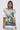 A woman with long dark hair and clear glasses stands against a gray background, wearing the NEW WORLD OVERSIZED GRAPHIC TEE with planetary and figure motifs, styled streetwear over a white lace skirt.