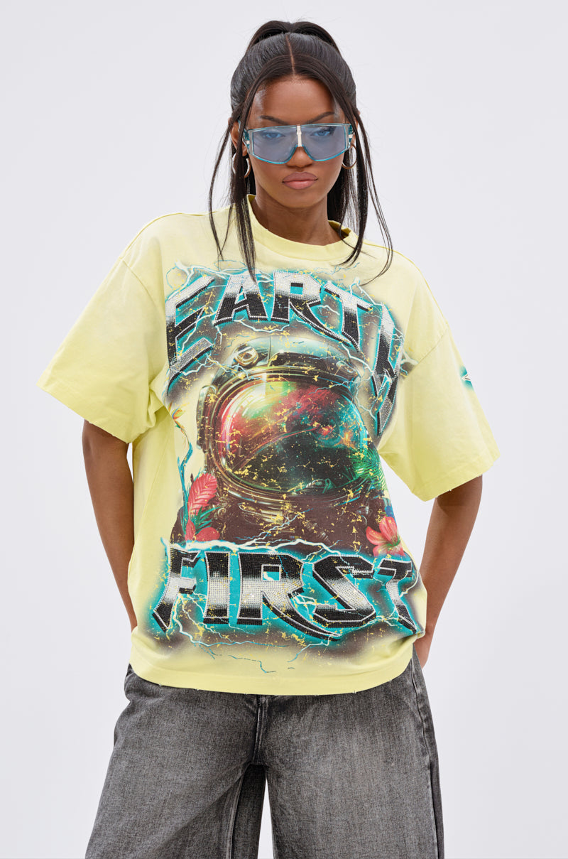 A woman wears blue-tinted sunglasses, hoop earrings, and the EARTH FIRST OVERSIZED GRAPHIC TEE featuring a colorful astronaut and "EARTH FIRST" text, styled with gray wide-leg jeans—ideal for fans of unisex streetwear and eco fashion.