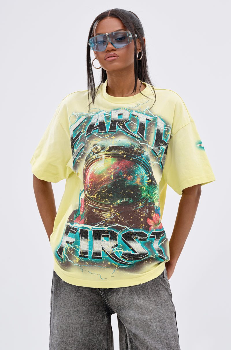 A woman models the EARTH FIRST OVERSIZED GRAPHIC TEE with a bold astronaut graphic and “EARTH FIRST” text, styled with sunglasses, hoop earrings, and gray jeans for a cool unisex streetwear look against a plain light background.