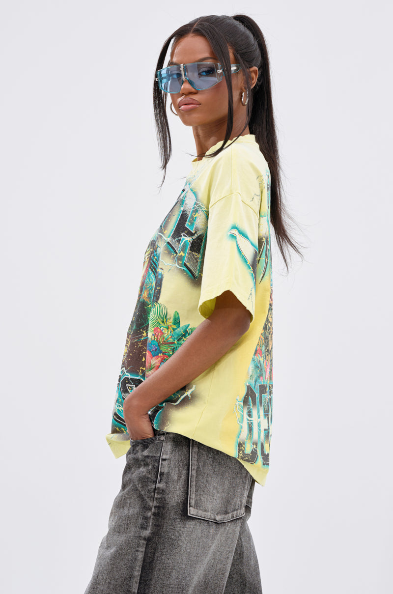 A woman with long dark hair in a high ponytail wears blue sunglasses, gray jeans, and the EARTH FIRST OVERSIZED GRAPHIC TEE—showcasing unisex streetwear style—standing hands-in-pockets against a plain light background.