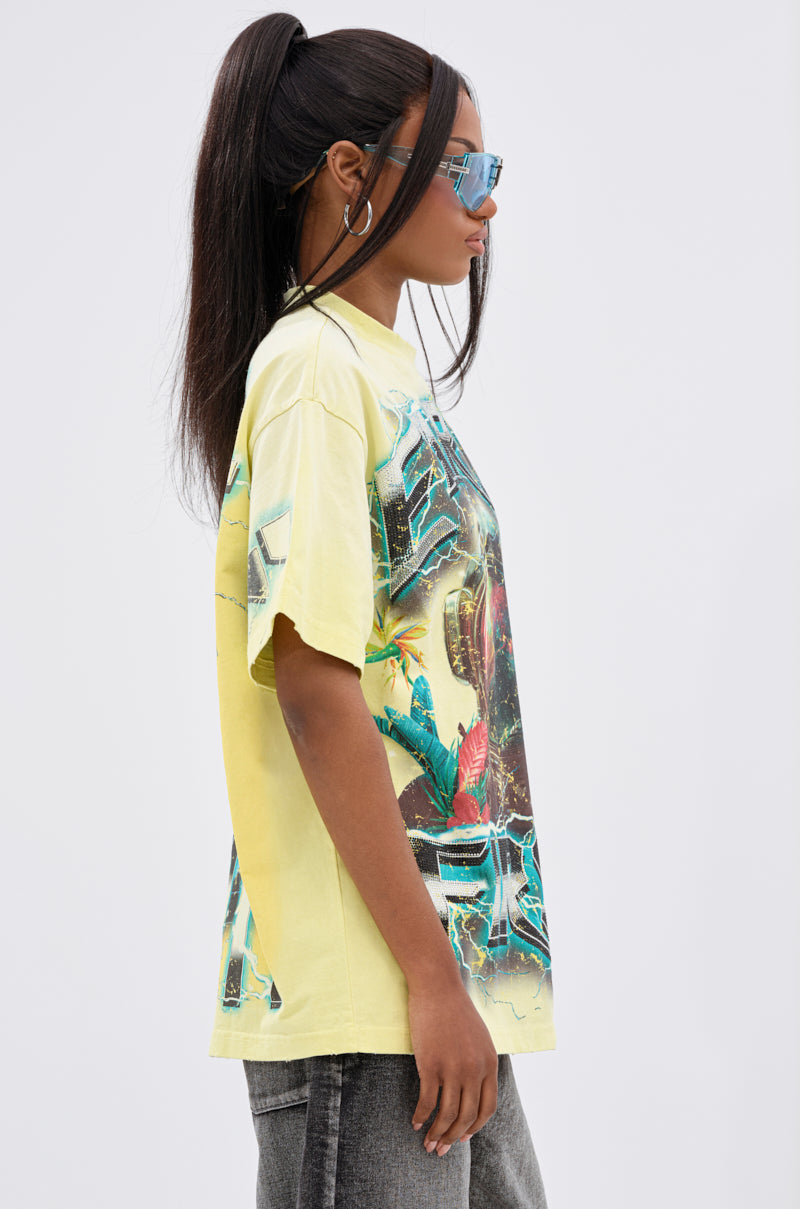 A woman with long dark hair in a ponytail wears blue sunglasses, gray jeans, and the EARTH FIRST OVERSIZED GRAPHIC TEE as she stands in profile against a plain light background.