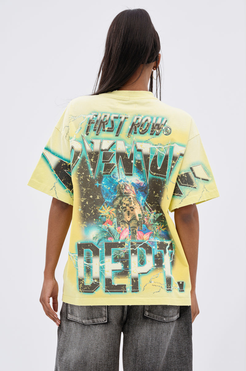 A person with long dark hair, seen from behind, wears the EARTH FIRST OVERSIZED GRAPHIC TEE featuring bold “FIRST ROW VENOM DEPT.” text and a colorful graphic. The yellow shirt is styled with dark jeans for an eco-conscious fashion look.