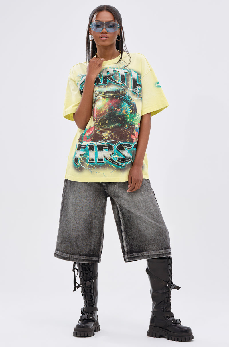 A person wearing the EARTH FIRST OVERSIZED GRAPHIC TEE, long gray denim shorts, tall black lace-up boots, and blue-tinted sunglasses stands against a plain light background.