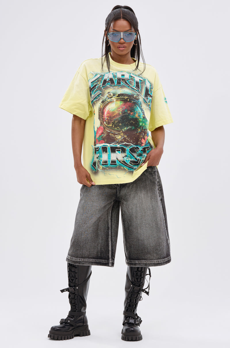Wearing blue sunglasses, the model pairs long black denim shorts and black lace-up boots with the EARTH FIRST OVERSIZED GRAPHIC TEE—featuring a vibrant astronaut design—while standing confidently against a plain white background.