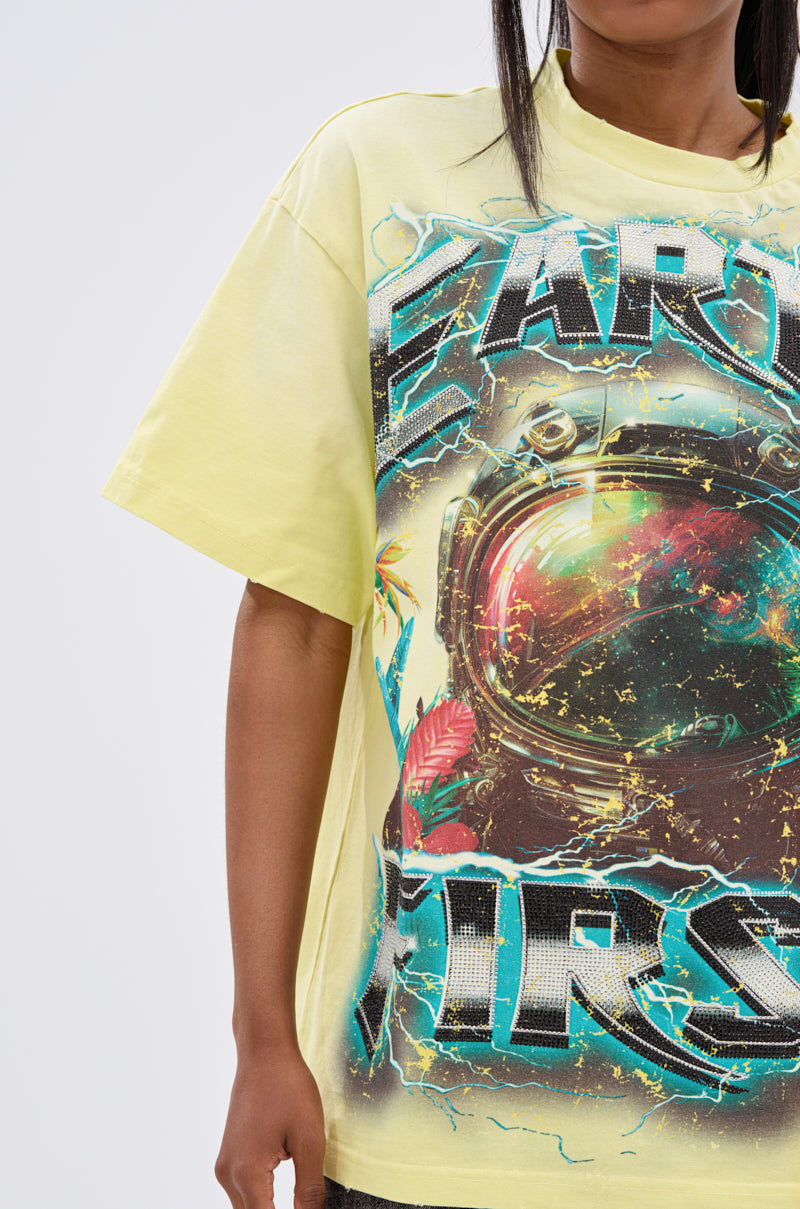 Wearing the EARTH FIRST OVERSIZED GRAPHIC TEE—a loose yellow shirt with a bold astronaut graphic—only the upper body and part of the arm are visible, highlighting eco-conscious fashion in a striking style.