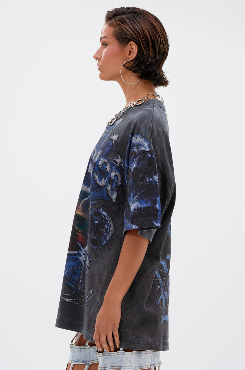 A person with short dark hair and large hoop earrings models the INSANE OVERSIZED TEE with ripped light-wash distressed cotton denim shorts, standing in profile against a plain white background.
