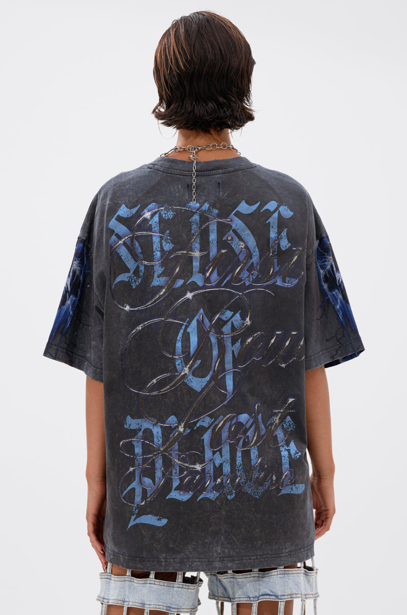 A person with short brown hair wears the INSANE OVERSIZED TEE—distressed black cotton, blue and white gothic "HOUSE OF BLUE" lettering on the back—paired with shredded denim shorts, against a plain light background.