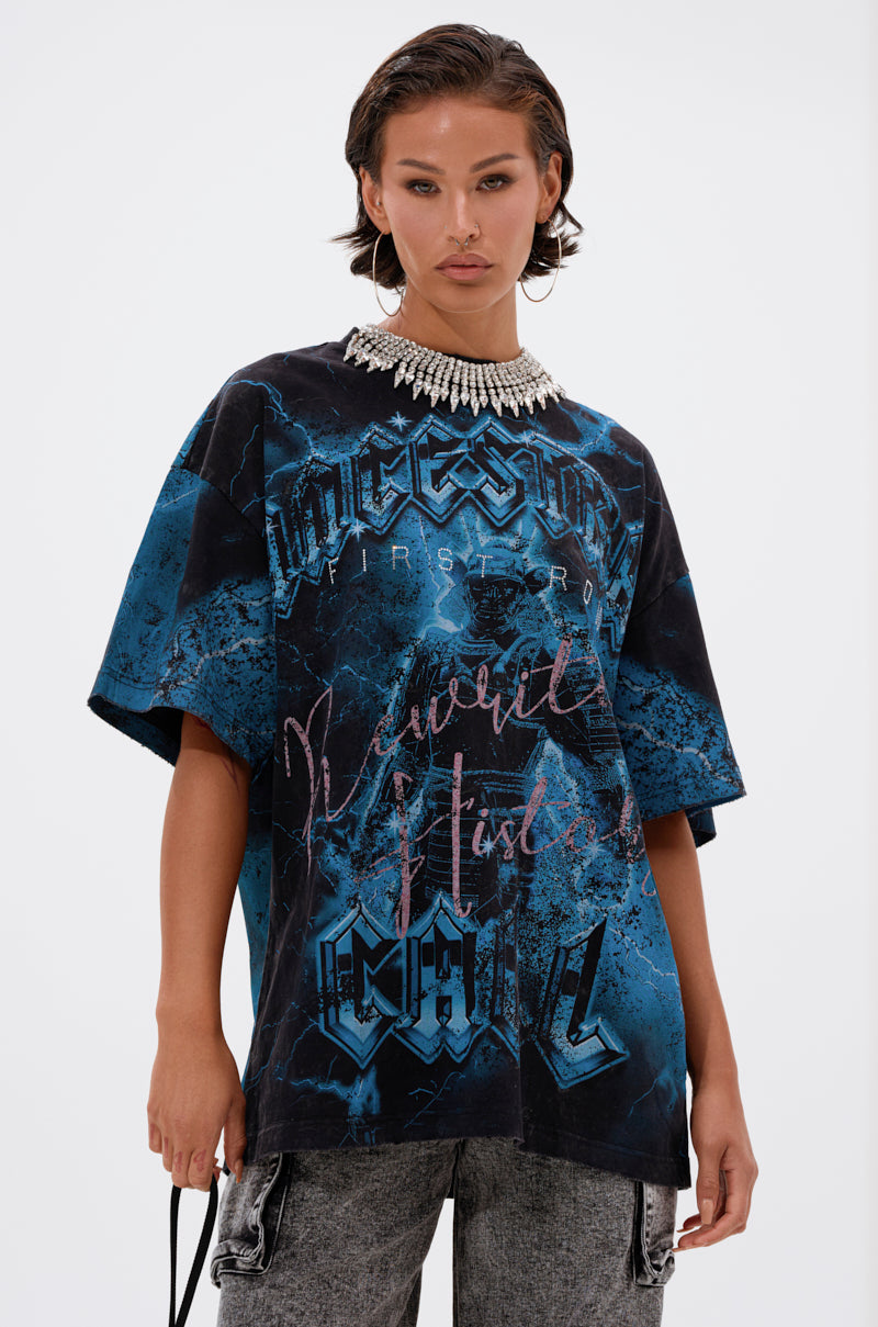 A woman with short dark hair wears a chunky pearl necklace, relaxed fit gray pants, and the ANCESTRAL FURY OVERSIZED GRAPHIC TEE in blue and black cotton with bold text, standing against a plain white background.