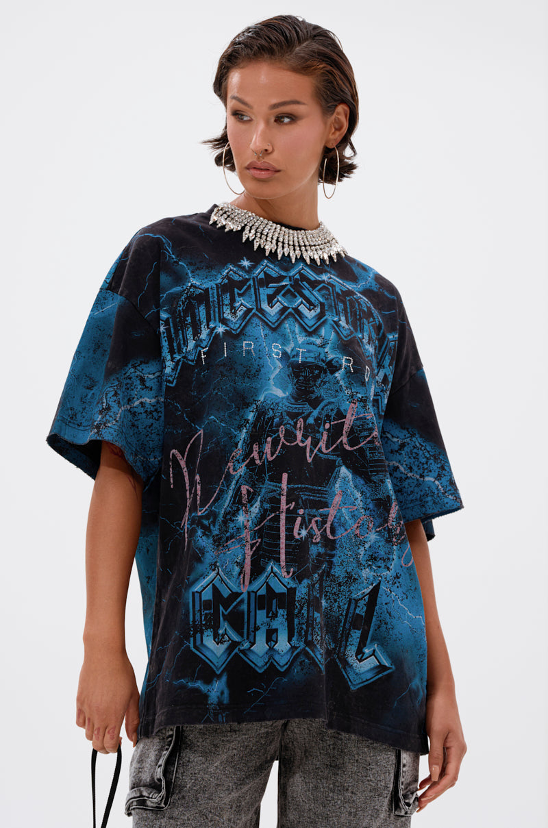 A woman with short dark hair wears the ANCESTRAL FURY OVERSIZED GRAPHIC TEE, featuring bold blue graphics, paired with gray jeans and a statement pearl necklace. She poses confidently against a plain white background, holding a small black strap.