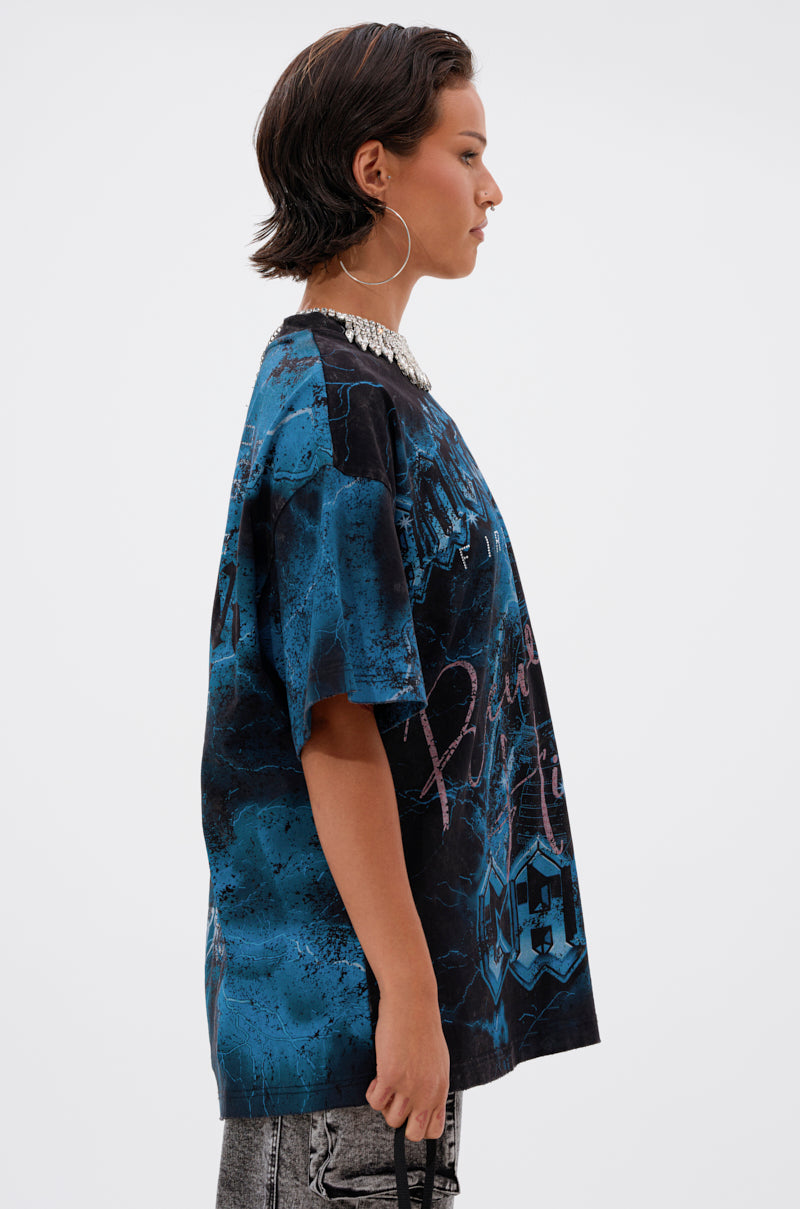 A person with short dark hair and large hoop earrings wears the ANCESTRAL FURY OVERSIZED GRAPHIC TEE in blue and black with a chunky necklace, standing in profile against a plain white background.