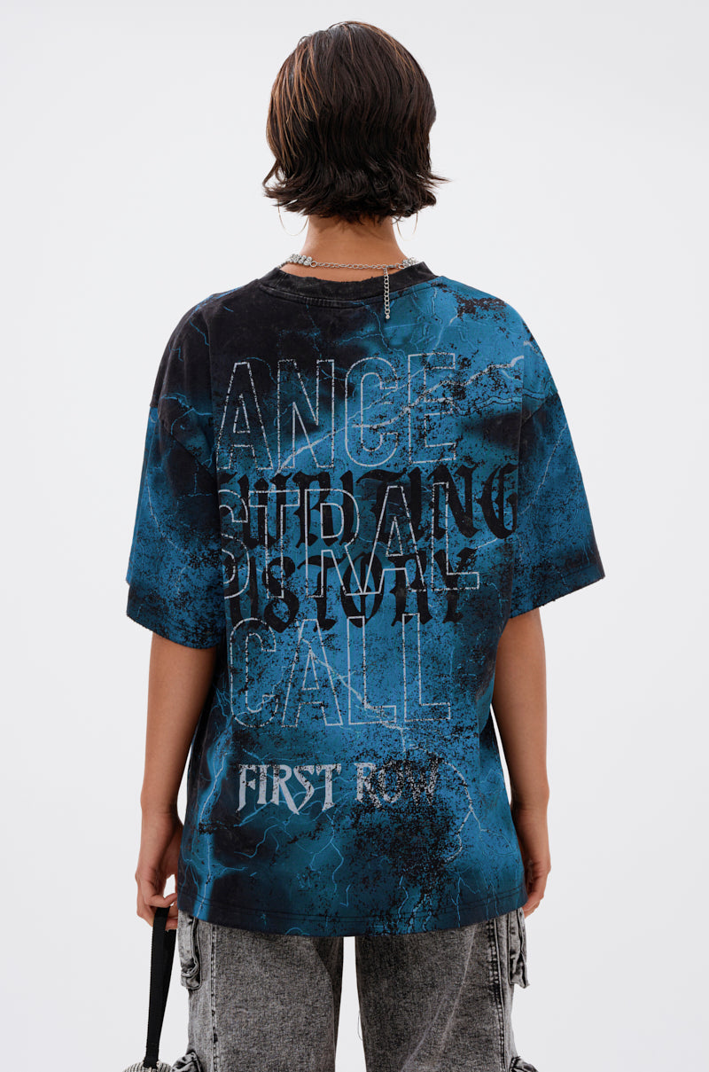 Seen from behind, a person with short, wavy brown hair wears the ANCESTRAL FURY OVERSIZED GRAPHIC TEE in blue and black featuring layered, textured graphics, paired with distressed gray jeans.
