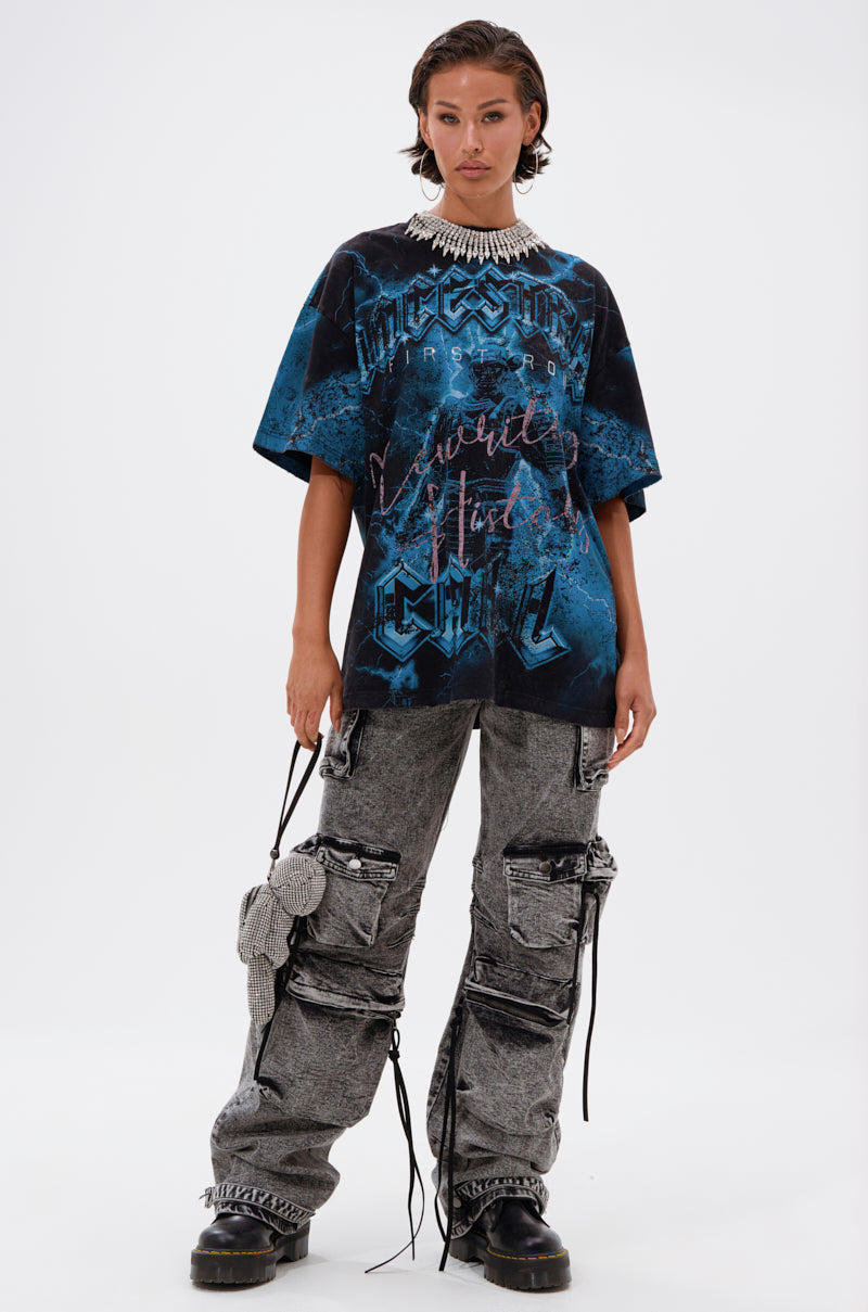 A person stands against a plain background wearing the ANCESTRAL FURY OVERSIZED GRAPHIC TEE, gray cargo pants with straps and pockets, chunky black boots, and a statement necklace. Their hair is short and slicked back.